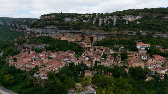 peaceful aerial view of a small village with stone buildings and clay roofs amidst cliffs and forests in a rural landscape with brown, red, green, and blue tones.