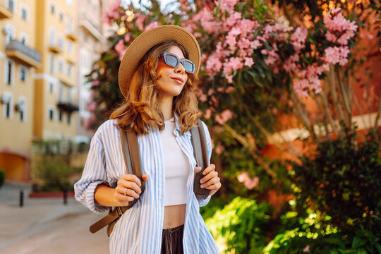 Young woman wearing hat and carrying backpack near blossoming trees on sunny day. Woman in sunglasses admires city's architecture. Lifestyle. Concepts of tourism, vacation, and rest.