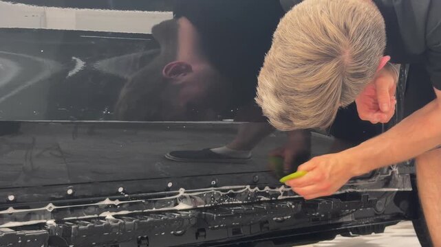 Professional car wrap installer applying a glossy transparent polyurethane paint protection film PPF on a vehicle body panel inside a premium auto detailing studio workshop.

