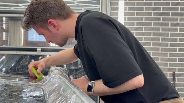 Professional car wrap installer applying a glossy transparent polyurethane paint protection film PPF on a vehicle body panel inside a premium auto detailing studio workshop.

