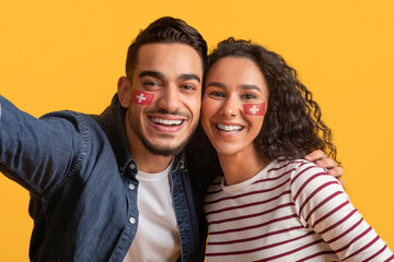 Fototapeta premium Smiling Multiracial Young Couple With Face Paint Celebrating At Studio With Yellow Background. Happy Emotion Displayed While Taking Selfie Together. Joyful Moment To Capture At Party.