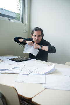 Angry middle-aged male teacher or office worker ripping paperwork at a desk covered in scattered documents, vertical shot