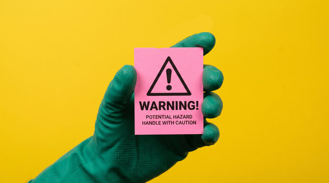 Hand wearing a green rubber glove holds a pink sticky note featuring a black warning symbol and text against a vibrant solid yellow studio background