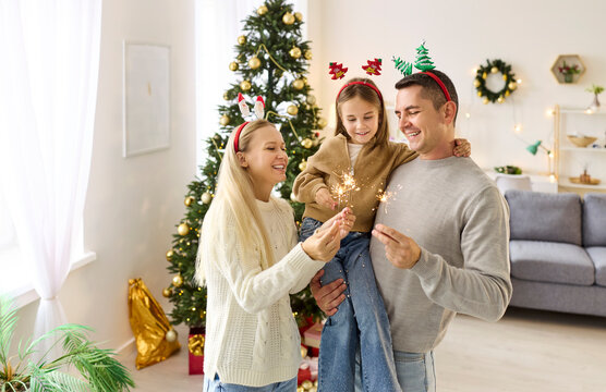 Happy family with child celebrate Christmas party with sparklers at home. Cheerful father holding daughter in arms by Xmas tree, people looking together at glowing lights with smiles, love and cuddle