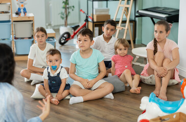 Obraz premium Boys and girls children sits on floor and listens to babysitter, organized pastime for children on summer holidays, kindergarten or day care activity
