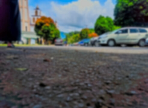 A blurred and defocused scene of a sunny outdoor car park with parked cars, green trees and towering buildings against a bright blue sky dotted with clouds. Viewed from a low angle on gravel ground.