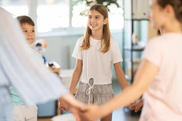 Young boys and girls of different ages stand in circle with young woman teacher, holding hands and moving together in playful ring dance © JackF