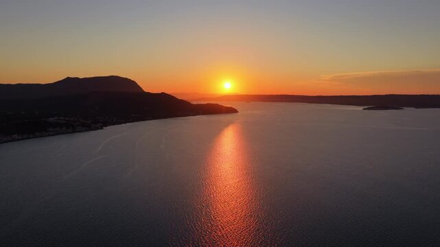 Aerial drone view of the sun setting into the narrow mouth of a large enclosed bay between dark headlands, Chania, Greece