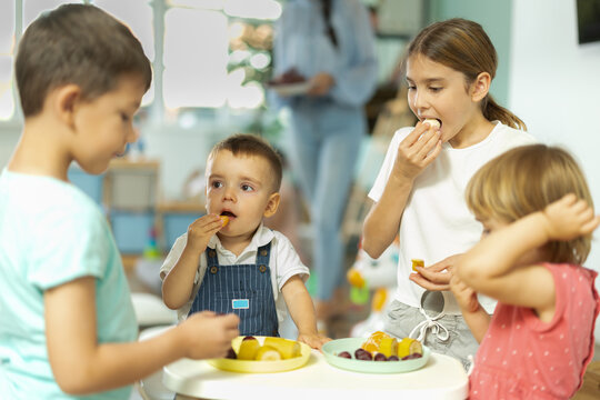 Young boys and girls sit at table enjoying fresh fruit including grapes, tangerines, and bananas while young woman teacher serves them