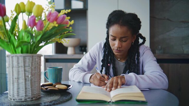 Young woman writes in notebook from book, while sitting at table with flowers and cookies in modern kitchen in morning light