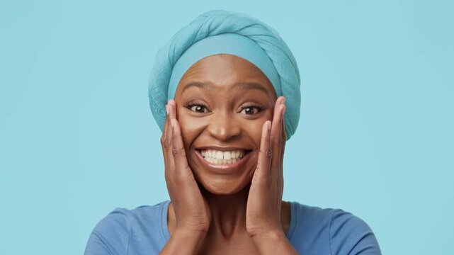 Joyful Emotion. Excited Black Woman Showing Enthusiasm With Hands On Face While Smiling In Studio Setting. Happy Expression Against Soft Blue Background. Positive Vibe And Celebration Concept.