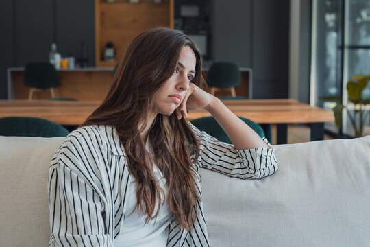 Woman feeling pensive, concerned, and apathetic, sitting alone on a couch indoors, contemplating her thoughts and experiencing loneliness during the day
