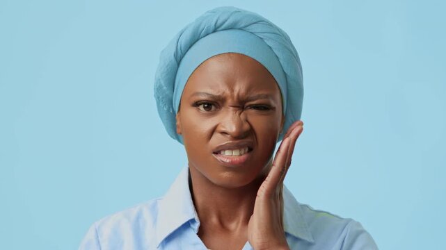 Worried Black Woman Touching Jaw While Experiencing Pain At Home, Displaying Emotion In Front Of Light Blue Background. Highlighting Importance Of Dental Health And Oral Care Concept.