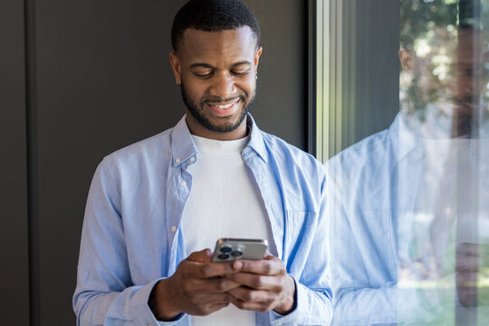 African american man smiling while holding a smartphone with both hands, typing on its screen and interacting with mobile technology, standing indoors next to a window showing his reflection