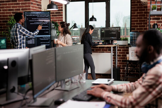 Technology team leader points out a source code mistake while software developers discuss an interface bug and slow data processing near a vertical screen.