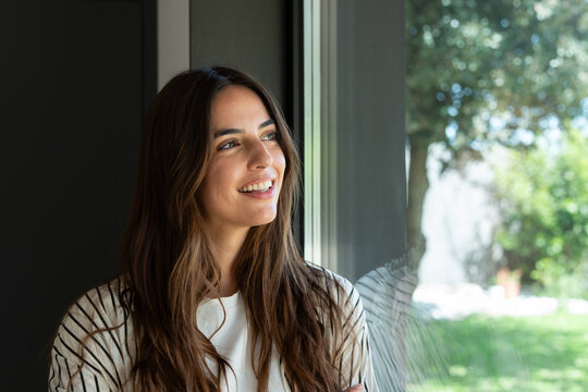 A smiling young woman with long brown hair gazes out a sunlit window in her home, appearing relaxed, hopeful and reflective. Natural light, casual clothing and a green backyard create a peaceful mood.