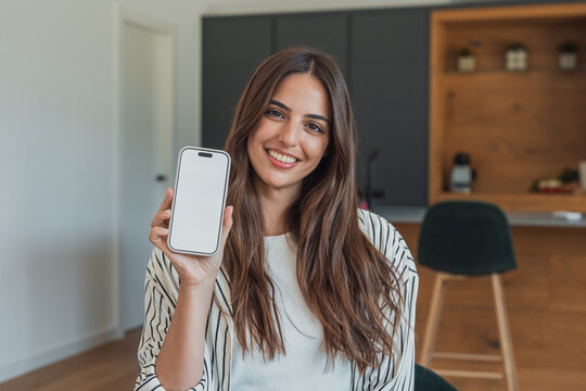 Smiling young woman in a modern kitchen holds up a smartphone with a blank screen, conveying friendly lifestyle, technology use, remote work and personal connection in a cozy home setting.