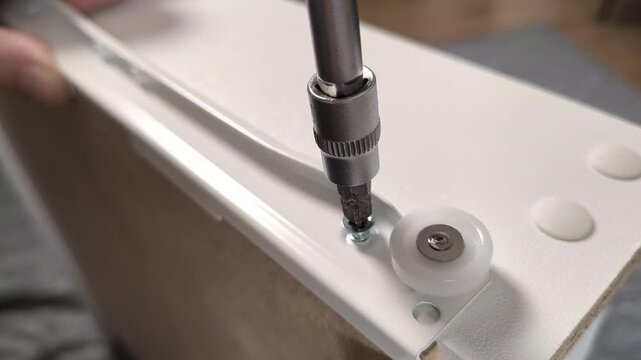Close-up of assembling furniture with a screwdriver tightening a screw near a roller wheel on a white panel, showcasing DIY installation, repair, and craftsmanship