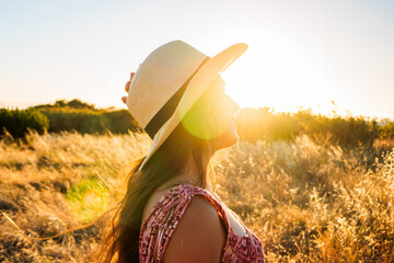 A young woman wearing a straw sun hat stands in a golden dry field, facing the warm sunset with a relaxed expression, backlit by lens flare that evokes summer warmth and peaceful solitude. © Daniel