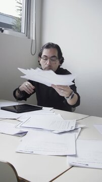 Vertical shot of a tired male teacher taking off his glasses and walking away from a desk cluttered with paperwork
