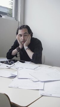 Vertical portrait of an exhausted male teacher sitting at a messy desk and rubbing his face in frustration