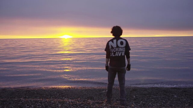  man on the beach at sunrise, captured from behind, wearing a black t-shirt with bold white text that reads "NO FUCKING SLAVE"