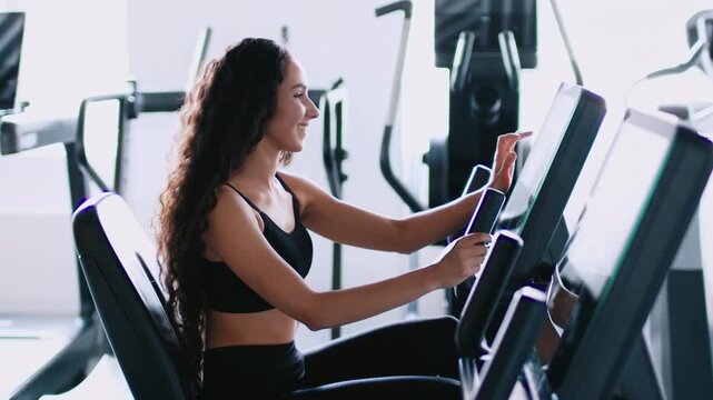 Smiling Young Latina Woman Engaged In Exercise While Using Elliptical Machine At Modern Gym. Focused On Working Out, She Represents Health And Fitness Lifestyle In The Gym Setting.