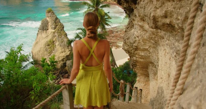 Female traveler standing on cliffside stairs looking at tropical ocean and beach bay