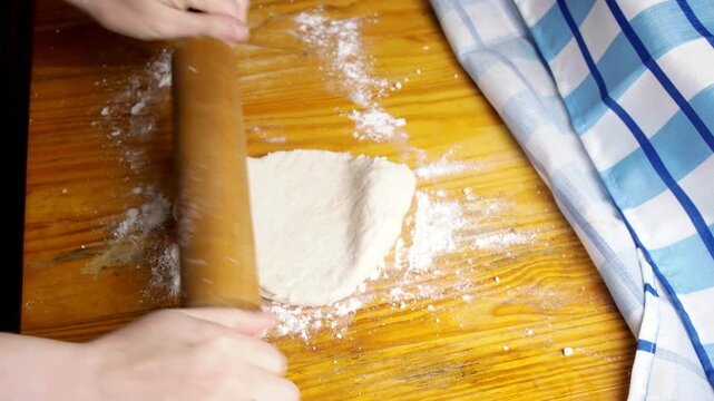 Top view of baker flattening raw dough with a wooden rolling pin on a floured table