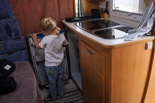  blonde girl kneeling on the floor and looking inside an open mini-fridge in a campervan kitchen.
