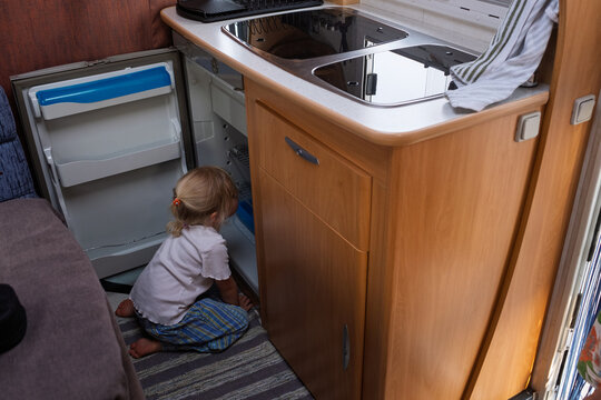 blonde girl kneeling on the floor and looking inside an open mini-fridge in a campervan kitchen.