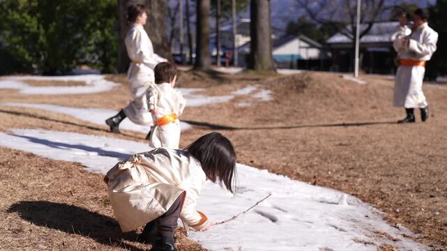 Children in white robes play with sticks in snowy yard. One child crouches, reaching for ground, joyful and active. Another child runs, arms outstretched, full of energy and laughter