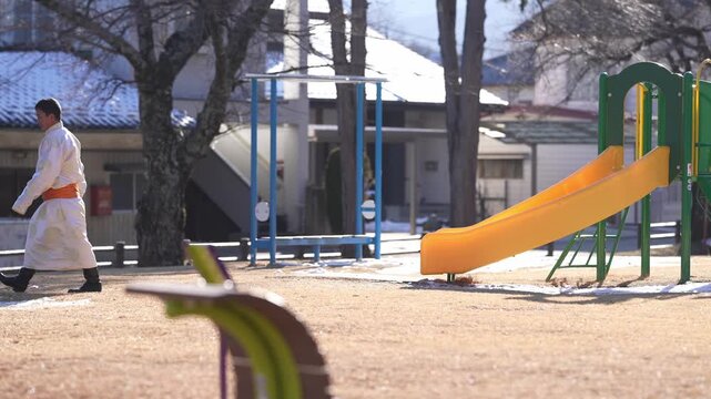 Bright sun bathes a quiet park playground. Man in white walks beside a small child. They pass a yellow slide and green structures. The ground is dry earth, trees stand bare