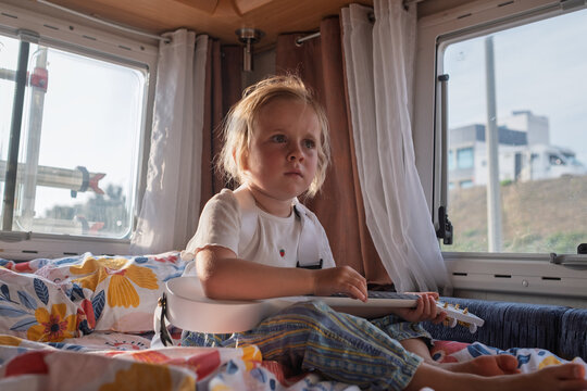 Cool little girl sitting cross-legged on a bed and playing a white ukulele in a sunlit campervan.