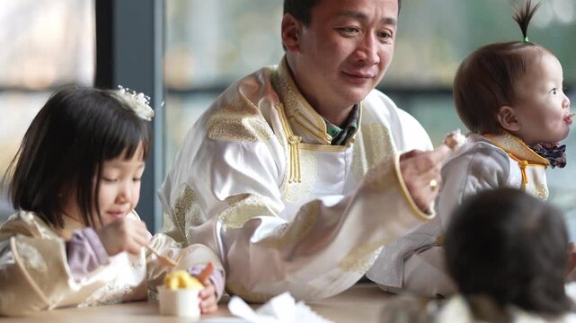 Man smiles while feeding two children at table. Children wear matching traditional outfits. Soft natural light fills the room. Heartwarming family moment captured. Ideal for cultural, parenting