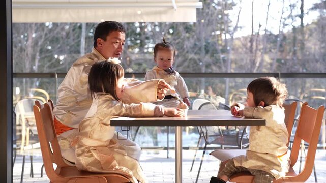 Warm light bathes a family dining together. Four children, one baby, share a meal with their dad. They smile, laugh, and eat with joy and comfort. The modern cafe offers a peaceful, sunlit setting