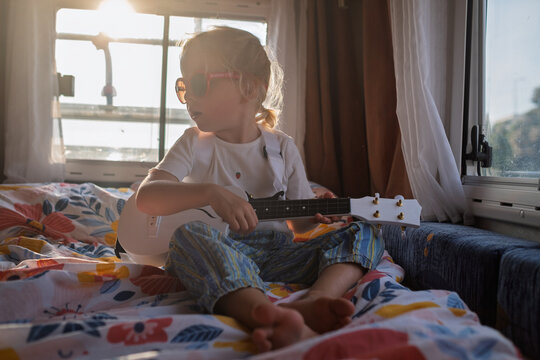 Cool little girl sitting cross-legged on a bed and playing a white ukulele in a sunlit campervan.