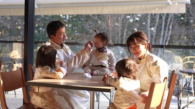 Warm sunlight bathes a cozy cafe setting. Family of five shares joyful moments. Parents smile, children laugh, hands touch. Soft chairs, wooden table, large windows