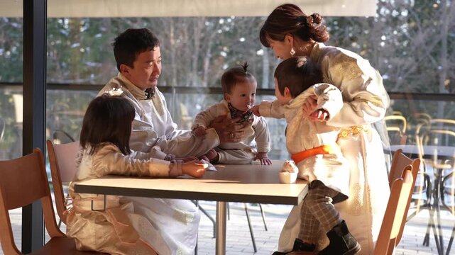Warm light streams through large windows. Family gathers around a table, joyful. Children laugh, play, and reach for food. Parents smile, embrace, and engage