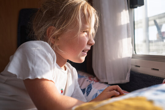  little blonde girl lying on a cozy bed inside a campervan and looking out the bright sunlit window.