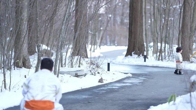 Snow blankets forest paths, tall trees stand silent. Man in white walks slowly, eyes forward. Child in matching attire skips ahead, joyful. Calm winter day, quiet forest, gentle movement