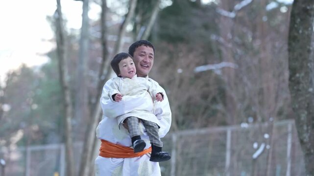 Man kneels in snow, holding child. He wears traditional winter robes. Child smiles, wrapped in white. Cold winter wind, quiet street. Perfect for family, cultural, or seasonal scenes