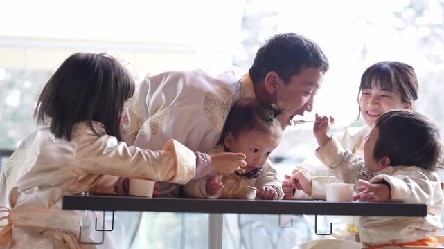 Family shares joyful mealtime together. Young children laugh and reach for food with parents nearby. Soft daylight illuminates their smiling faces. Cute scene captures tender family moments