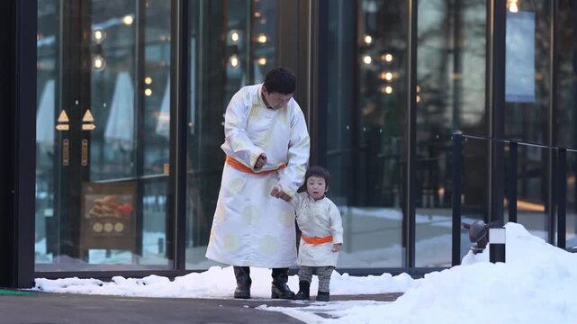 Man and child walk through snow outside building. They wear matching white robes with orange belts. Child holds man's hand as they step carefully. Snow covers ground near glass entrance doors