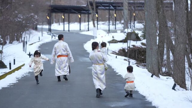 Winter path lined with snow and bare trees. Family dressed in white robes with orange sashes. Children run joyfully, adults walk calmly behind. Snowy path leads to wooden covered walkway