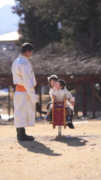 Warm sunlight bathes a rustic outdoor scene. Man in white stands beside a wooden ride. Child smiles, seated on a red toy. Another child sits behind, holding on tight
