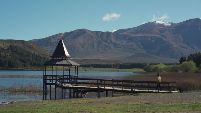 Man wearing a yellow jacket walking on a wooden pier extending into Laguna La Zeta, with a gazebo and mountains in the background, Esquel, Chubut, Argentina.