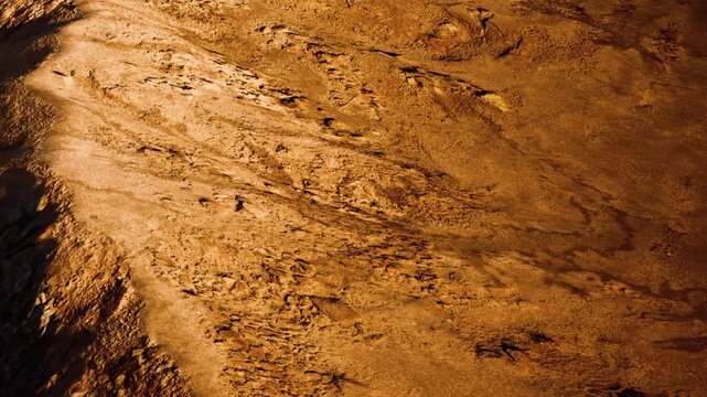 The ground shows a detailed texture with various patterns made by rocks and sand. The lighting highlights the surface shapes. This landscape appears dry and barren.