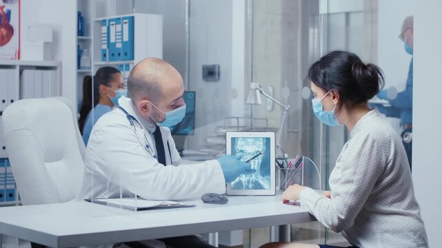 Woman patient reviews an X ray on a digital tablet through a plexiglass wall, showing protected diagnosis and medical consultation.