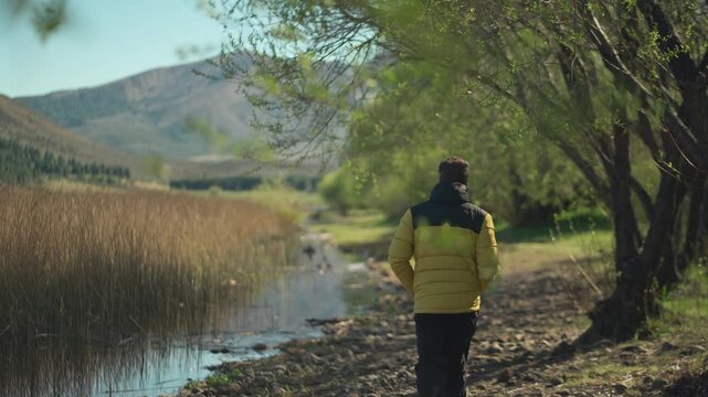 Man in a yellow jacket walking along a dirt trail through a lush, green woodland next to a calm lake, outdoor lifestyle scene.